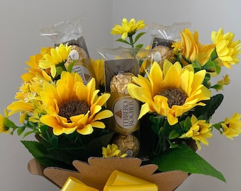 May include: A decorative bouquet featuring bright yellow sunflowers and smaller yellow flowers, arranged in a brown cardboard container. The bouquet includes Ferrero Rocher chocolates and is adorned with a large yellow bow.