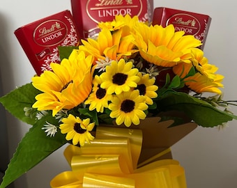 May include: A bouquet of yellow sunflowers with a red and gold bow. The bouquet is arranged in a brown paper bag. There are three boxes of Lindt Lindor chocolates in the bouquet.