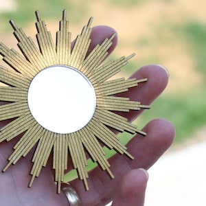 May include: A gold sunburst mirror with a round, reflective surface. The mirror is made of wood and has a rustic, vintage look.
