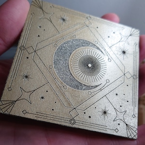 May include: A gold square tile with a black and white crescent moon design. The moon is surrounded by a geometric pattern of lines and dots. The tile has a textured surface.