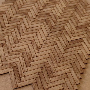 May include: Close-up of a wooden parquet floor tile sample. The tile features a herringbone pattern with varying shades of brown and tan, mimicking the look of natural wood. The sample is on a light brown surface.