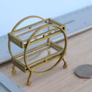 May include: A miniature gold-coloured serving cart with two glass shelves and small wheels. The cart has a rounded design with a coin next to it for scale. The cart is on a light wood surface.