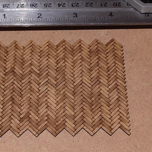May include: A rectangular piece of miniature parquet flooring, constructed from small, light brown wooden blocks arranged in a herringbone pattern. A ruler with inch measurements is visible in the background, indicating the scale of the flooring.