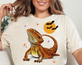 May include: Beige t-shirt featuring a detailed illustration of a bearded dragon in shades of orange and brown. The design includes a full moon, bats, and stars, creating a Halloween theme. The shirt is a casual wear item.
