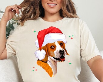 May include: A beige t-shirt with a beagle dog wearing a red and white Santa hat. The dog is looking to the right and has a happy expression. There are green and red glitter accents around the dog.