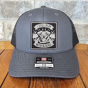 May include: A gray and black trucker hat with a patch that says "I Still Play With Blocks". The patch features a stylized image of a motorcycle engine. The hat is adjustable and has a Richardson 112 label.