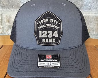 May include: A gray and black trucker hat featuring a black shield-shaped patch on the front. The patch displays the text YOUR CITY FIRE/RESCUE, the number 1234, and the word NAME. A sticker on the brim reads RICHARDSON ORIGINAL 112 TRUCKER.