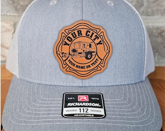 May include: A gray trucker hat with a white mesh back and a leather patch. The patch features a fire truck illustration and the text "YOUR CITY" and "YOUR NAME OR TEXT". The hat is adjustable and has a Richardson label.