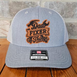 May include: Gray and white trucker hat with a brown leather patch that says "Dad Fixer of Things" with a hammer and wrench design. The hat has a black adjustable strap with a Richardson 112 logo.