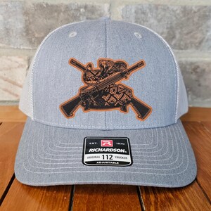 May include: A gray and white trucker hat with a brown leather patch featuring a snake and crossed rifles. The patch has the text "EST. 1970 RICHARDSON. ORIGINAL TRUCKER 112 ADJUSTABLE".