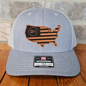 May include: Gray trucker hat with a white mesh back and a leather patch in the shape of the United States. The patch features a flag design with the text "2ND". The hat has a curved brim and an adjustable strap.