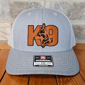 May include: A gray and white trucker hat with a brown leather patch that says "K9" with a black silhouette of a dog's head inside the "K". The hat has a Richardson 112 adjustable label.
