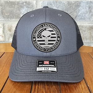 May include: A gray and black trucker hat with a black and white skull graphic and the text "Second Amendment Defend Yourself". The hat has a red and white label with the text "Richardson 112 Original Trucker Adjustable".