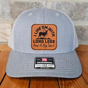 May include: A gray and white trucker hat with a brown leather patch that says "I Like 'Em With Long Legs And A Big Rack" with a silhouette of a deer with antlers. The patch has a target symbol and a deer silhouette. The hat has a Richardson 112 label.