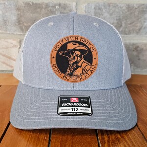 May include: Gray trucker hat with a white mesh back and a circular patch featuring a skull wearing a cowboy hat. The patch reads "DO IT WITH GRIT OR DONT BOTHER AT ALL." The hat has a Richardson tag that reads "ORIGINAL 112 TRUCKER ADJUSTABLE."