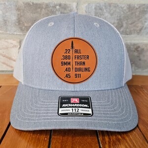 May include: A gray and white trucker hat with a brown leather patch that reads ".22 ALL .380 FASTER 9MM THAN .40 DIALING .45 911". The hat is adjustable and has a Richardson logo on the back.