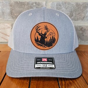 May include: A gray and white trucker hat with a brown leather patch featuring a silhouette of a deer in a forest setting. The patch has the text "EST. 1970 RICHARDSON. ORIGINAL TRUCKER 112 ADJUSTABLE" on it.