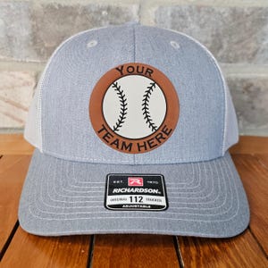 May include: Gray and white trucker hat with a brown leather patch featuring a baseball and the text "YOUR TEAM HERE". The hat is adjustable and has a Richardson logo.