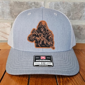 May include: Gray trucker hat with a white mesh back and a patch featuring a hooded skull holding a rifle. The patch is brown and orange. The hat has a curved brim and an adjustable strap. The label reads "RICHARDSON ORIGINAL 112 TRUCKER ADJUSTABLE".