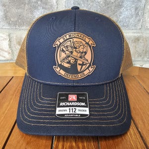 May include: A navy blue and brown trucker hat with a brown leather patch featuring a winged angel holding a sword and shield with the text "St. Michael Defend Us". The hat has a brown mesh back and a black label with the text "Richardson. Original 112 Trucker Adjustable" and a red "R" logo.