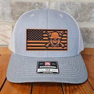 May include: A gray and white trucker hat with a brown leather patch featuring an American flag with a skull and crossbones design. The patch also includes the text "EST. 1970 RICHARDSON. ORIGINAL TRUCKER 112 ADJUSTABLE".