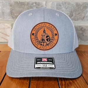 May include: A gray and white trucker hat with a brown leather patch featuring a Spartan warrior and the text "Molon Labe Come And Take It". The hat is adjustable and has the Richardson logo and "112" on the label.