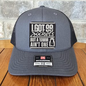May include: A gray and black trucker hat with a white patch that reads "I GOT 99 SOCKETS BUT A 10MM AIN'T ONE" with a small image of a 10mm socket. The hat has a black mesh back and a red and white label that reads "RICHARDSON ORIGINAL 112 TRUCKER ADJUSTABLE".