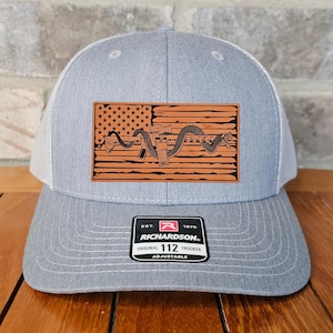 May include: A gray and white trucker hat with a brown leather patch featuring an American flag with a snake and a rifle. The patch also includes the text "EST. 1970 RICHARDSON. ORIGINAL TRUCKER 112 ADJUSTABLE".
