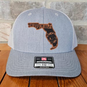 May include: A gray and white trucker hat with a brown leather patch featuring the outline of the state of Florida with the word "FIREFIGHTER" inside. The patch also includes a firefighter helmet graphic. The hat has a black label with the text "EST. 1970 RICHARDSON. ORIGINAL TRUCKER 112 ADJUSTABLE".