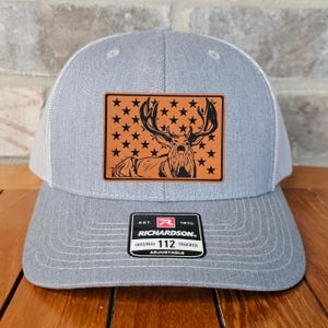 May include: A gray and white trucker hat with a brown leather patch featuring a black silhouette of a buck with large antlers in front of a field of stars. The patch has the text "EST. 1970 RICHARDSON. ORIGINAL TRUCKER 112 ADJUSTABLE".