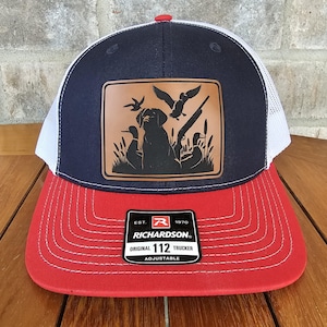 May include: A navy blue and red trucker hat with a brown leather patch featuring a silhouette of a dog and ducks. The patch also includes the text "EST. 1970 RICHARDSON. ORIGINAL 112 TRUCKER ADJUSTABLE".