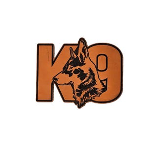 May include: A tan leather patch featuring the letters "K9" with a black silhouette of a dog's head. The dog is facing left, with its ear and eye visible. The patch has a black border.