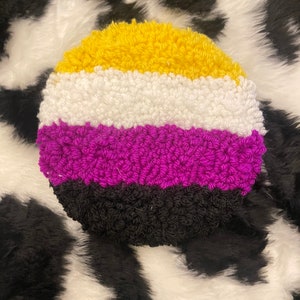 May include: A round, crocheted cushion featuring the colours of the non-binary pride flag: yellow, white, purple, and black.