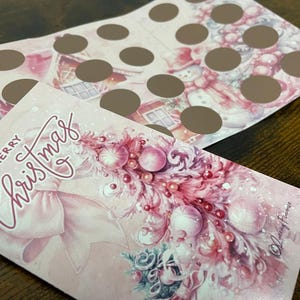 Pu&ograve; includere: Un biglietto di Natale rosa con un fiocco rosa e un albero di Natale rosa e bianco decorato con ornamenti. Il biglietto dice "Buon Natale" in scrittura rosa.