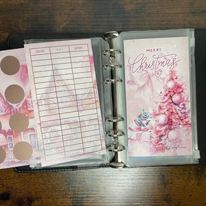 Pu&ograve; includere: Un planner natalizio rosa e bianco con una tasca trasparente sul davanti. La tasca contiene un biglietto con il testo "Buon Natale" e un'illustrazione rosa e bianca di un albero di Natale. Il planner ha un raccoglitore ad anelli argentato e una pagina con un motivo rosa e bianco con una griglia.
