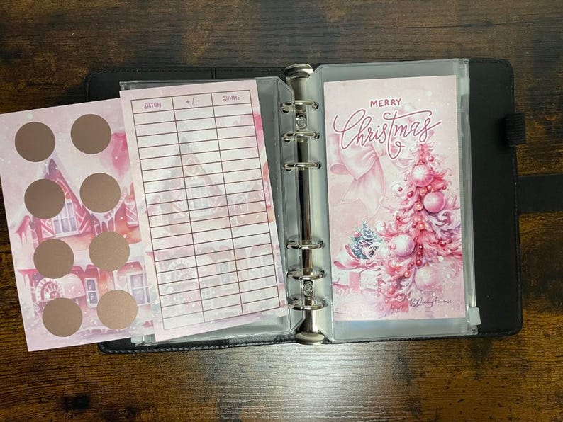 Pu&ograve; includere: Un planner natalizio rosa e bianco con una tasca trasparente sul davanti. La tasca contiene un biglietto con il testo "Buon Natale" e un'illustrazione rosa e bianca di un albero di Natale. Il planner ha un raccoglitore ad anelli argentato e una pagina con un motivo rosa e bianco con una griglia.