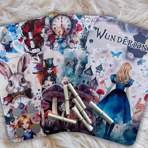 Könnte beinhalten: Drei rechteckige Planereinlagen mit Alice im Wunderland-Illustrationen, einem Kaninchen, Pilzen, einer Uhr und dem Text WUNDERLAND. Die Seiten haben seitliche Stanzlöcher und enthalten mehrere kleine, helle zylindrische Marker.