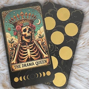 Könnte beinhalten: Eine Tarotkarte mit schwarzem Hintergrund und goldenen Akzenten. Die Karte zeigt ein Skelett, das eine Krone und Blumen trägt und sich die Ohren zuhält. Die Karte trägt den Titel "The Drama Queen".