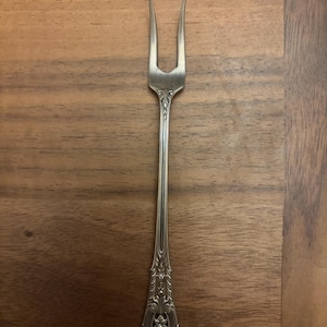 May include: A silver-toned metal cocktail fork featuring two long, slender tines and an elongated handle. The handle is decorated with intricate, embossed floral and scrollwork patterns that extend from the base up to the neck of the fork.