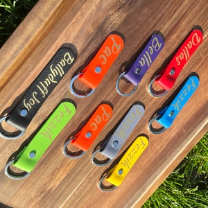 May include: Seven leather keychains with gold lettering on a wooden surface. The keychains are in various colors, including black, green, orange, purple, red, blue, and gray. The names on the keychains are "Baldyfoot Joy", "Fezzie", "Pac", "Fezzie", "Bella", "Dallas", and "Fezzie".