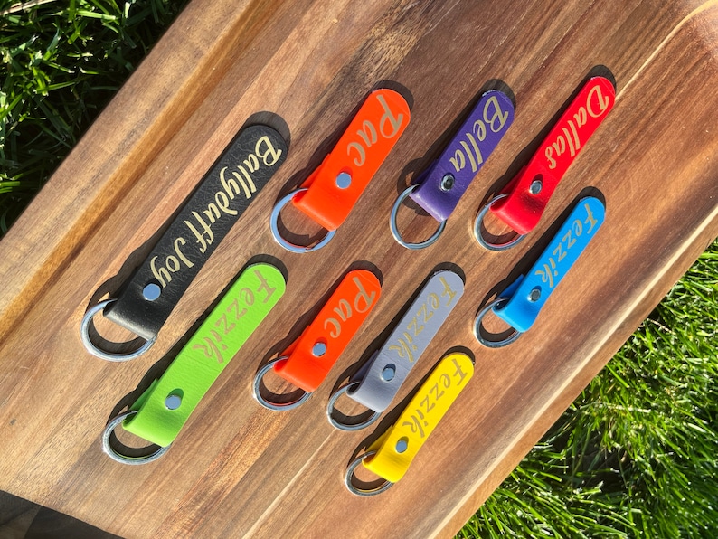 May include: Seven leather keychains with gold lettering on a wooden surface. The keychains are in various colors, including black, green, orange, purple, red, blue, and gray. The names on the keychains are "Baldyfoot Joy", "Fezzie", "Pac", "Fezzie", "Bella", "Dallas", and "Fezzie".