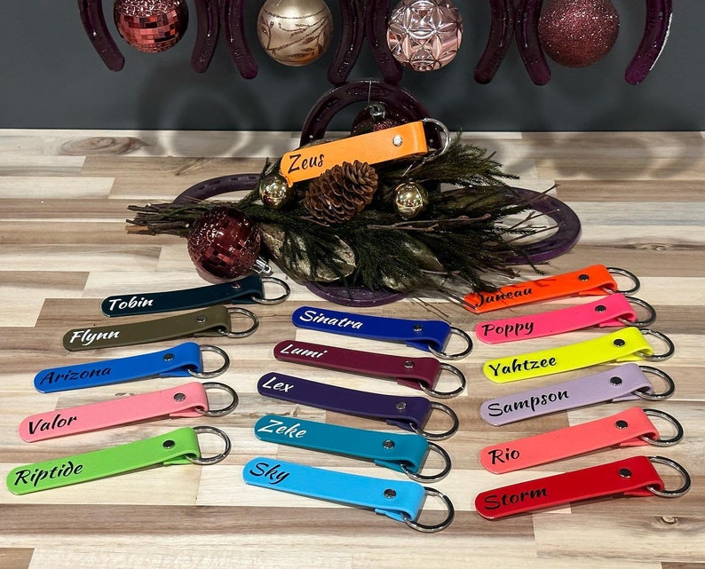 May include: A collection of 17 colorful leather keychains with personalized names, including "Zeus", "Tobin", "Flynn", "Arizona", "Valor", "Riptide", "Sinatra", "Lumi", "Lex", "Zeke", "Sky", "Janeau", "Poppy", "Yahtzee", "Sampson", "Rio", and "Storm".