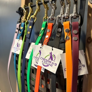 Puede incluir: Una variedad de correas para perros de colores con cierres metálicos colgados en un estante. Las correas están hechas de diferentes materiales, incluyendo cuero y caucho. Las correas están etiquetadas con el texto "Cesar K-9 & Equine".