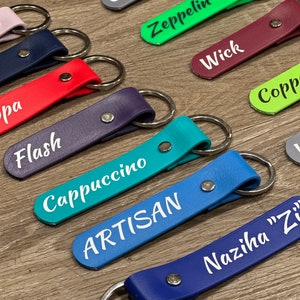 May include: A collection of colorful leather keychains with different names printed on them, including "Cowboy", "Zeppelin", "Wick", "Cappuccino", "Artisan", and "Naziha "Z"".