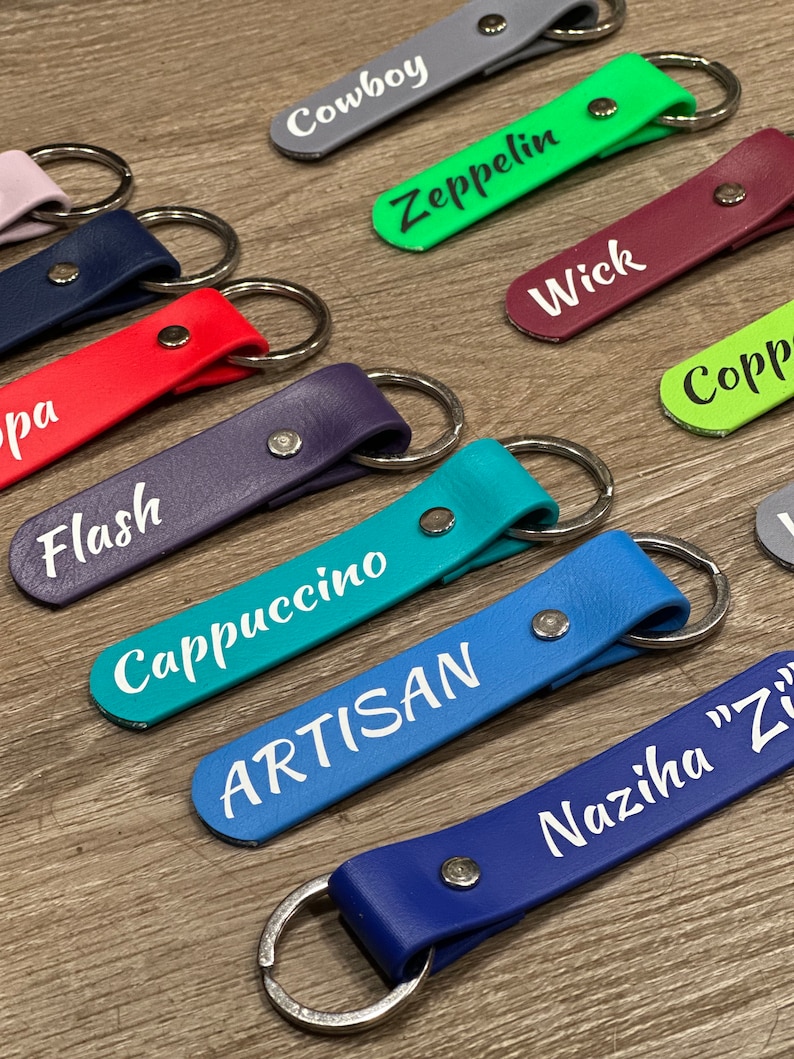 May include: A collection of colorful leather keychains with different names printed on them, including "Cowboy", "Zeppelin", "Wick", "Cappuccino", "Artisan", and "Naziha "Z"".