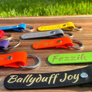 May include: A collection of colorful leather keychains with different names engraved in gold lettering. The keychains are on a wooden surface. The names on the keychains are "Fezzik", "Pac", and "Ballyduff Joy".