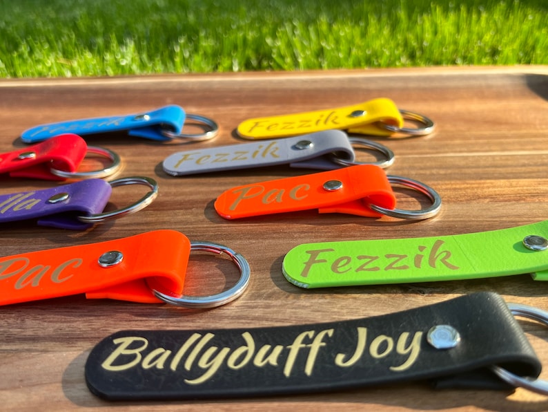 May include: A collection of colorful leather keychains with different names engraved in gold lettering. The keychains are on a wooden surface. The names on the keychains are "Fezzik", "Pac", and "Ballyduff Joy".