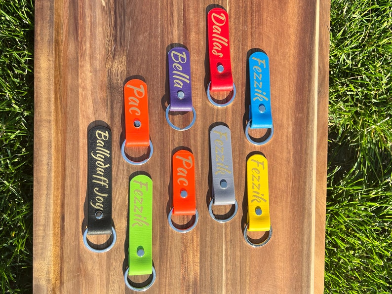 May include: Eight colorful leather keychains with gold lettering. The keychains are on a wooden surface with green grass in the background. The names on the keychains are: Dallas, Fezzik, Bella, Pac, Fezzik, Ballyduff Joyo, Pac, Fezzik.