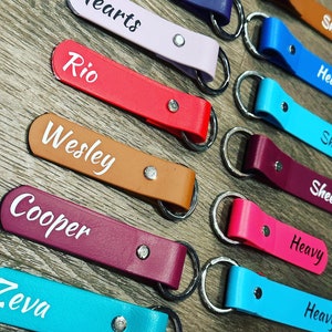 May include: A collection of personalized leather keychains with various names, including "Hearts", "Rio", "Wesley", "Cooper", "Zeva", "Heavy", and "Shea". The keychains are in different colors, including red, pink, brown, purple, blue, and turquoise.