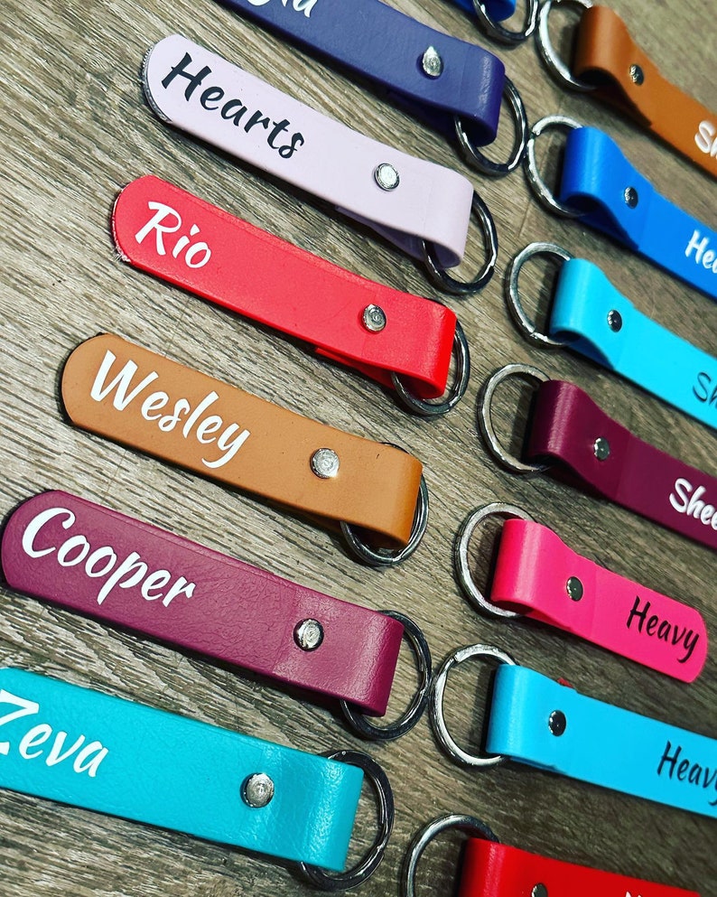 May include: A collection of personalized leather keychains with various names, including "Hearts", "Rio", "Wesley", "Cooper", "Zeva", "Heavy", and "Shea". The keychains are in different colors, including red, pink, brown, purple, blue, and turquoise.